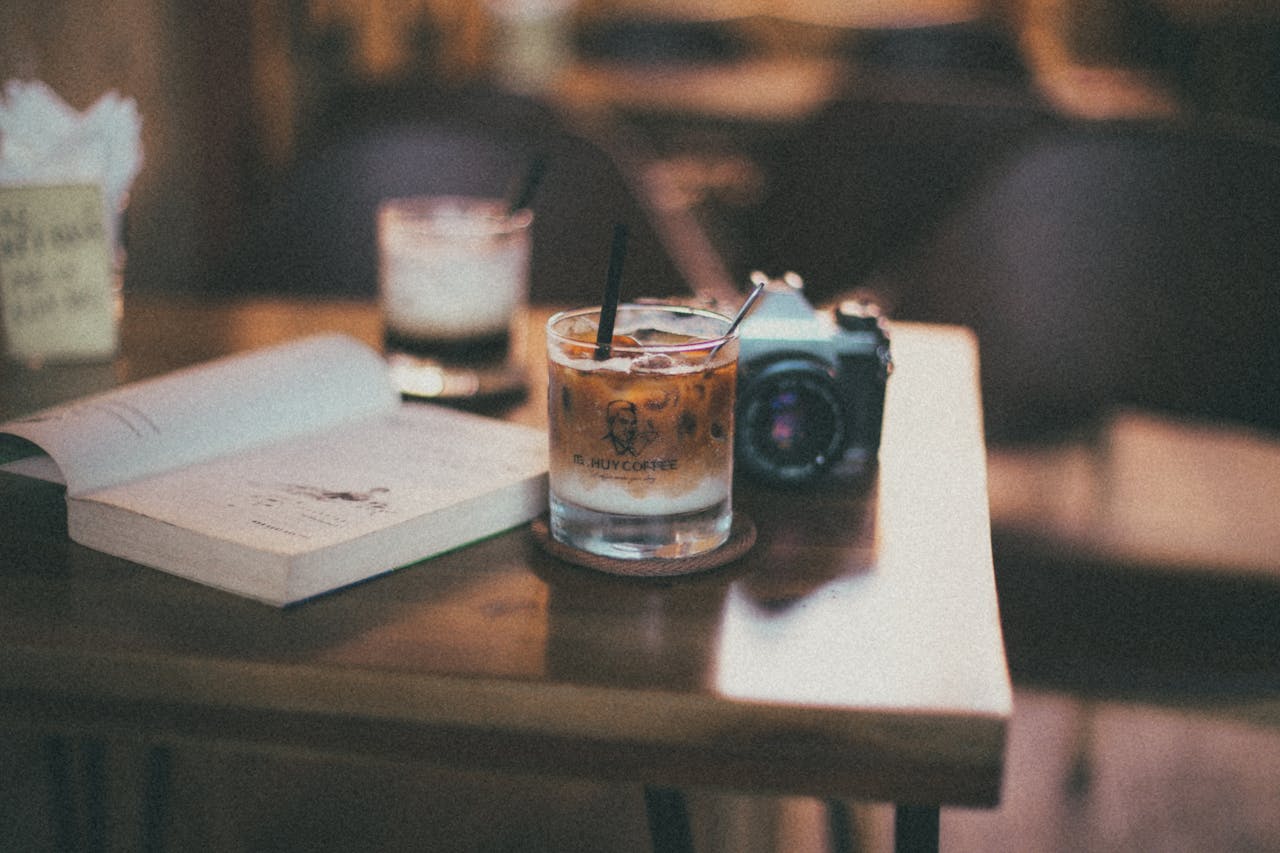 Warm cafe setting with a book, iced coffee, and camera on a wooden table.
