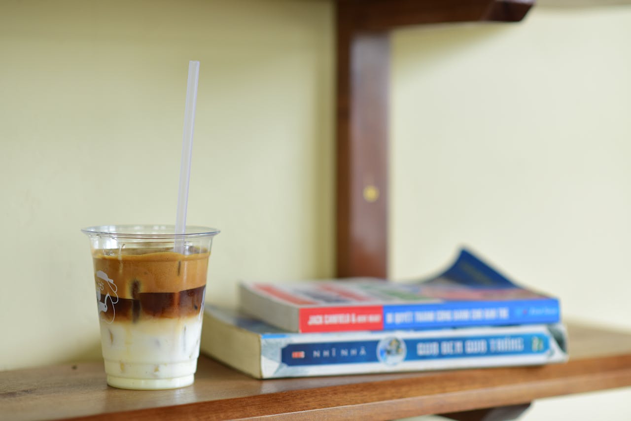A close-up of a refreshing iced latte in a plastic cup next to books on a wooden shelf.